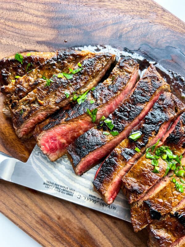 Sliced medium-rare steak with a seared crust, inspired by a classic Carne Asada recipe, garnished with chopped herbs and displayed on a wooden cutting board with a knife partially visible underneath.