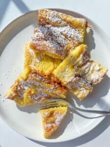 A plate of golden, fluffy baked French Toast Casserole pieces dusted with powdered sugar, with a fork resting on the plate and sunlight casting gentle shadows.