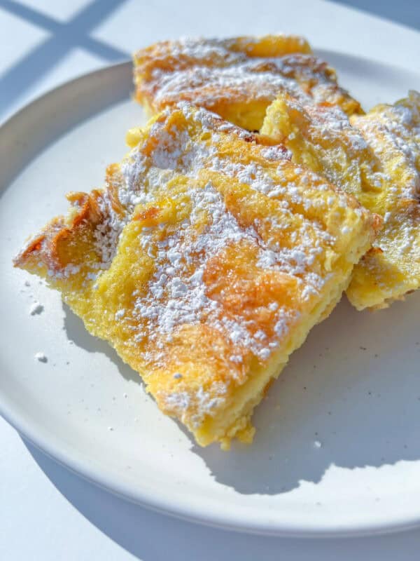 Three golden, fluffy slices of baked pancake, reminiscent of a French Toast Casserole, are dusted with powdered sugar and arranged on a white plate, bathed in natural sunlight.
