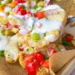 A close-up of a fork lifting loaded cauliflower nachos topped with diced red tomatoes, green onions, red onions, melted cheese, and a drizzle of white sauce on a parchment-lined tray.