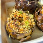 Close-up of Italian stuffed artichokes in a baking dish, topped with golden breadcrumbs and fresh green herbs. Sunlight highlights the crispy texture and browned edges.