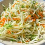 A close-up of a bowl filled with fresh coleslaw made of shredded cabbage, carrots, and parsley in a light dressing. This Healthy Coleslaw Recipe is bright and colorful, served in a beige bowl on a white surface.