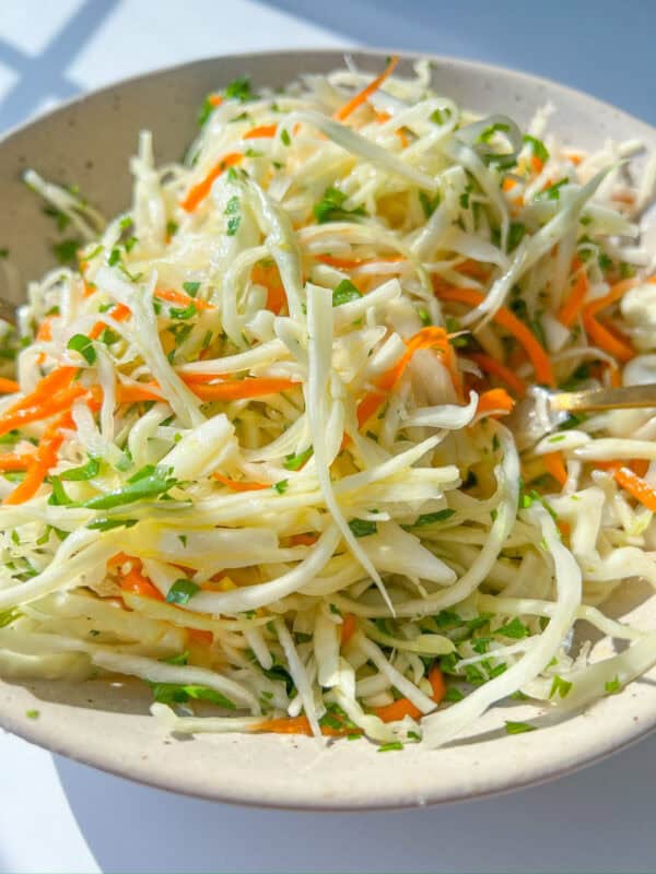 A close-up of a bowl filled with fresh coleslaw made of shredded cabbage, carrots, and parsley in a light dressing. This Healthy Coleslaw Recipe is bright and colorful, served in a beige bowl on a white surface.