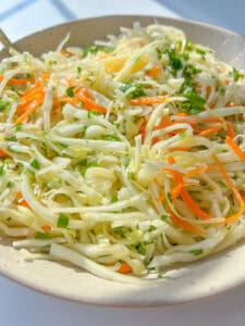 A close-up of a healthy coleslaw recipe featuring thinly sliced cabbage, carrots, and chopped herbs in a white bowl.