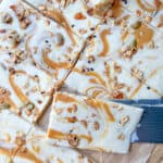 A close-up of Cottage Cheese Bark dessert bars with a creamy white topping swirled with caramel and sprinkled with granola, sitting on parchment paper. A knife is partially placed under one of the bars.