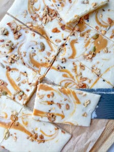 A close-up of Cottage Cheese Bark dessert bars with a creamy white topping swirled with caramel and sprinkled with granola, sitting on parchment paper. A knife is partially placed under one of the bars.