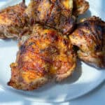 Four pieces of Grilled Chicken Thighs with crispy, golden-brown skin and char marks are arranged on a white plate, bathed in bright natural sunlight.