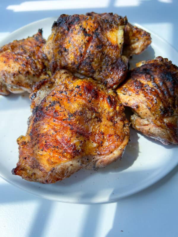 Four pieces of Grilled Chicken Thighs with crispy, golden-brown skin and char marks are arranged on a white plate, bathed in bright natural sunlight.
