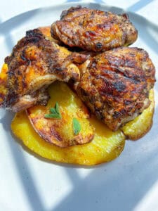Three pieces of Grilled Chicken Thighs are served atop roasted golden potato slices and garnished with a small sprig of fresh herbs, all beautifully arranged on a white plate.