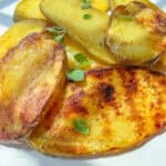 Sliced grilled potatoes with golden, crispy edges, garnished with fresh green herbs, served on a white plate in natural sunlight.