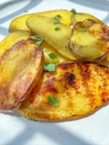 Sliced grilled potatoes with golden, crispy edges, garnished with fresh green herbs, served on a white plate in natural sunlight.