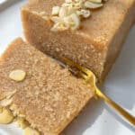 A close-up of a rectangular slice of Semolina Cake topped with sliced almonds on a white plate. A gold fork rests beside the cake, with one piece of the Semolina Cake partially cut and ready to eat.