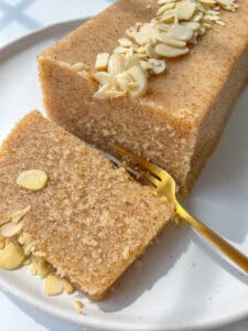 A close-up of a rectangular slice of Semolina Cake topped with sliced almonds on a white plate. A gold fork rests beside the cake, with one piece of the Semolina Cake partially cut and ready to eat.