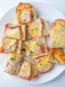 Slices of bread topped with ham, melted cheese, and chopped herbs are arranged on a white plate. These Bulgarian Meat Sandwiches (Prinzesi) are cut into small rectangles, with the cheese lightly browned for extra flavor.