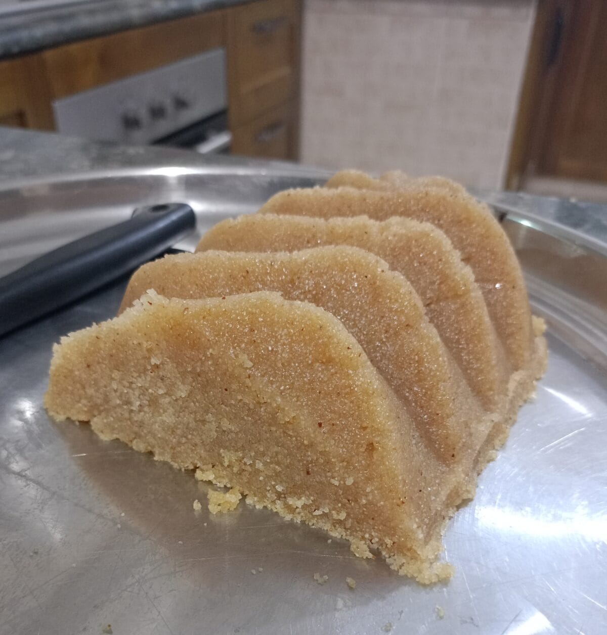 A piece of semolina halwa dessert, shaped with ridges, sits on a metal plate in a kitchen with wooden cabinets and an oven in the background.