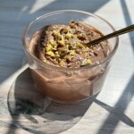 A glass cup filled with creamy healthy chocolate mousse topped with chopped nuts, with a spoon inside, sits on a sunlit wooden surface.