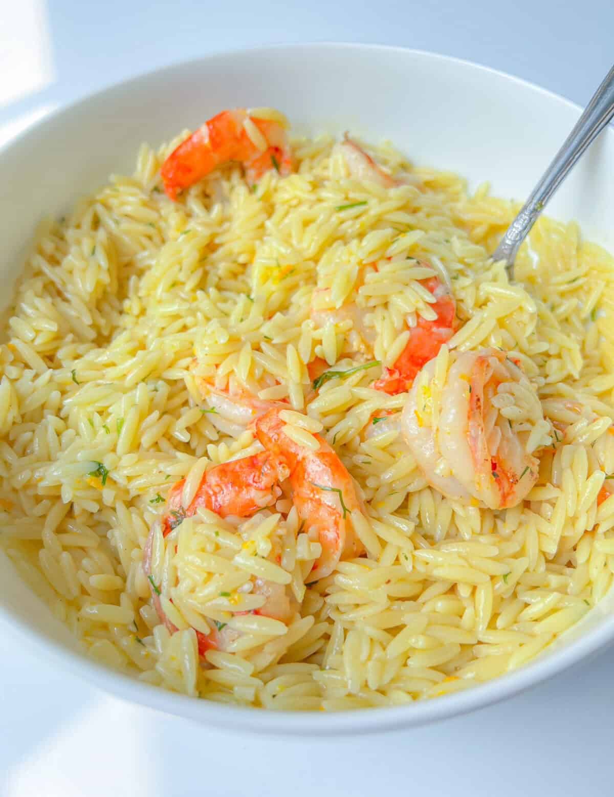 A white bowl filled with creamy Lemon Feta Orzo pasta and several cooked shrimp, with a fork resting inside. The dish has a light, glossy appearance and is set on a white surface.