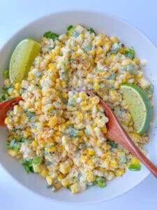A white bowl filled with creamy Mexican Corn Salad, garnished with chopped green herbs, two lime wedges, and sprinkled with chili powder. Two wooden serving spoons are resting in the salad.