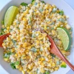 A bowl of creamy Mexican Corn Salad with chopped green peppers and herbs, garnished with two lime wedges and sprinkled with spices. Two wooden spoons rest in the bowl.