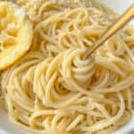 A close-up of Pasta Limone features spaghetti topped with grated cheese in a white bowl, a gold fork elegantly twirling the noodles, and a squeezed lemon half resting on the side.