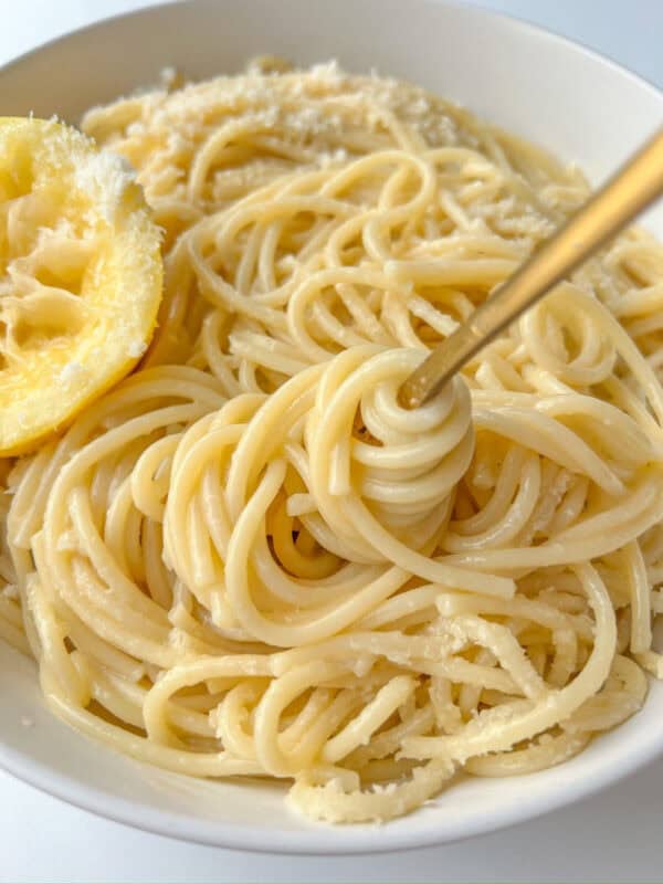 A close-up of Pasta Limone features spaghetti topped with grated cheese in a white bowl, a gold fork elegantly twirling the noodles, and a squeezed lemon half resting on the side.