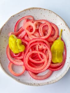 A ceramic plate with spiral-cut pickled red onions from a classic Pickled Onions Recipe and two yellow pepperoncini peppers on a white background.