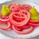 A plate with thinly sliced pickled red onions, inspired by a classic Pickled Onions Recipe, and two yellow pepperoncini peppers on a speckled white dish against a white background.
