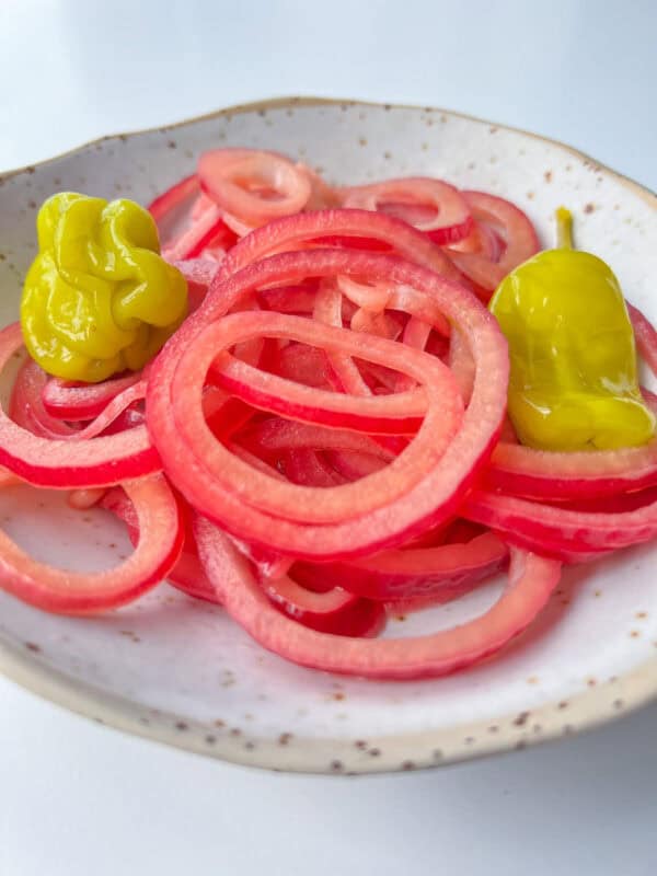 A plate with thinly sliced pickled red onions, inspired by a classic Pickled Onions Recipe, and two yellow pepperoncini peppers on a speckled white dish against a white background.