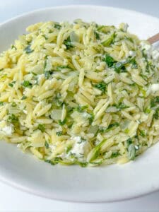 A white plate filled with Spinach Orzo Salad, featuring orzo pasta mixed with chopped herbs, crumbled feta cheese, finely diced onions, and fresh spinach. The dish appears creamy and vibrant.