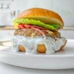 A close-up of a burger on a white plate. The burger features a creamy white sauce dripping down, a patty, tomato slices, and fresh lettuce, all sandwiched in a toasted bun. The background is a blurred kitchen setting.