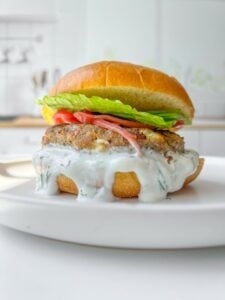 A close-up of a burger on a white plate. The burger features a creamy white sauce dripping down, a patty, tomato slices, and fresh lettuce, all sandwiched in a toasted bun. The background is a blurred kitchen setting.