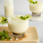 A creamy lime drink garnished with lime zest and mint sits on a wooden tray with lime wedges, mint, and ice cubes; another drink and a bottle of the beverage are in the background—perfect alongside Big Mac Smash Tacos.