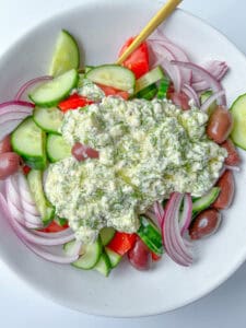 A bowl of Greek Feta Salad with sliced cucumbers, tomatoes, red onions, Kalamata olives, and a generous dollop of tzatziki sauce on top, with a spoon resting in the salad.