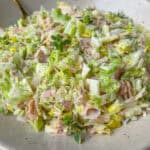 A close-up of a bowl filled with chopped cabbage, ham slices, and herbs, lightly seasoned with black pepper. A spoon rests in the bowl, showcasing this creamy and fresh Italian Chopped Salad.