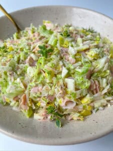 A close-up of a bowl filled with chopped cabbage, ham slices, and herbs, lightly seasoned with black pepper. A spoon rests in the bowl, showcasing this creamy and fresh Italian Chopped Salad.