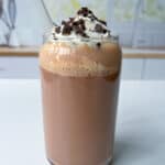 A glass of iced Nutella Chocolate Milk topped with whipped cream and chocolate shavings, served with a clear straw, set on a white surface with a blurred kitchen background.