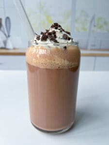A glass of iced Nutella Chocolate Milk topped with whipped cream and chocolate shavings, served with a clear straw, set on a white surface with a blurred kitchen background.