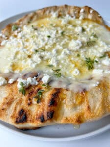 A close-up of a Honey and Feta Pita, featuring grilled flatbread topped with melted cheese, crumbled feta, and fresh herbs, served on a white plate.