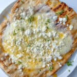 A Honey and Feta Pita—grilled flatbread topped with melted cheese, crumbled white feta, fresh green herbs, and a drizzle of honey—is served on a white plate.