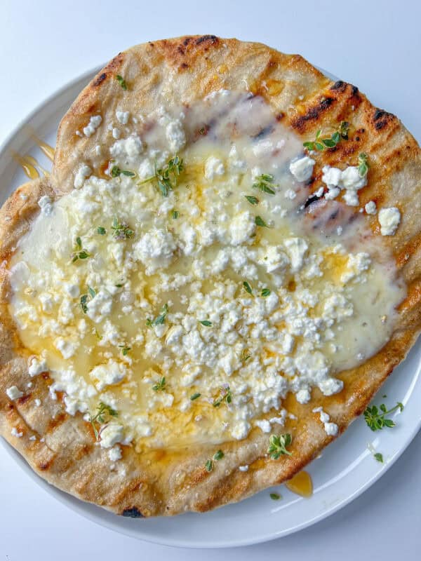 A Honey and Feta Pita—grilled flatbread topped with melted cheese, crumbled white feta, fresh green herbs, and a drizzle of honey—is served on a white plate.