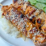 Two Teriyaki Salmon Skewers topped with sesame seeds, served over white rice with sliced cucumbers on a white plate.