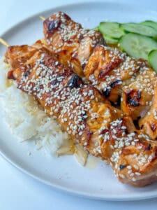 Two Teriyaki Salmon Skewers topped with sesame seeds, served over white rice with sliced cucumbers on a white plate.