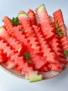 A bowl filled with crinkle-cut watermelon fries, garnished with small green herb sprigs, on a light background—making for a refreshing and fun watermelon snack.