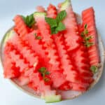 A plate of crinkle-cut Watermelon Fries garnished with fresh mint and thyme leaves, set on a light background, makes for a refreshing watermelon snack.