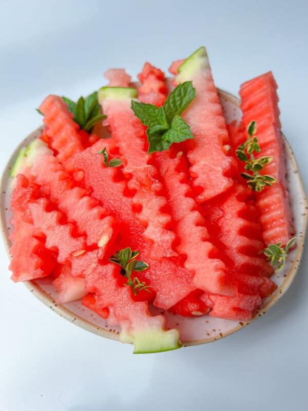 A plate of crinkle-cut Watermelon Fries garnished with fresh mint and thyme leaves, set on a light background, makes for a refreshing watermelon snack.