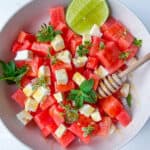 A refreshing Watermelon Feta Salad featuring cubed watermelon, feta cheese, fresh mint, thyme, a drizzle of honey, and half a lime on the side. A honey dipper rests on top for added sweetness.