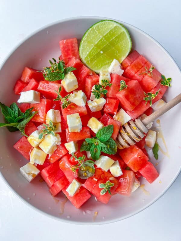 A refreshing Watermelon Feta Salad featuring cubed watermelon, feta cheese, fresh mint, thyme, a drizzle of honey, and half a lime on the side. A honey dipper rests on top for added sweetness.