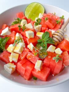 A vibrant bowl of Watermelon Feta Salad, featuring cubed watermelon and feta cheese, garnished with fresh mint and herbs, a honey dipper with honey, and a half lime.