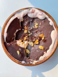 A wooden bowl filled with strawberry yogurt topped with a layer of dark chocolate and sprinkled with chopped walnuts. The chocolate is cracked, revealing the creamy Strawberry Yogurt Bowl beneath.