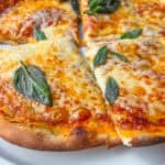 A close-up of an Easy Homemade Pizza topped with fresh basil leaves, cut into slices on a white plate with a pizza cutter nearby.